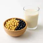 신선한 노란 콩과 검은콩이 담긴 목재 그릇과 두유 한 잔이 정갈하게 놓인 장면입니다.