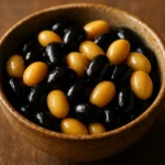 따뜻하고 윤기가 흐르는 조리된 검은콩과 대두가 나무 테이블 위 도자기 그릇에 정갈하게 담긴 모습입니다.